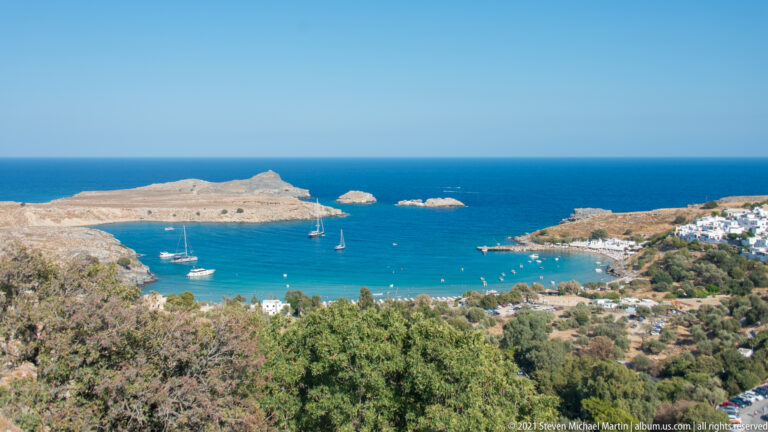 Turquoise sheltered bay near Lindos with anchored sailboats, rocky headlands, and white buildings along the shore.