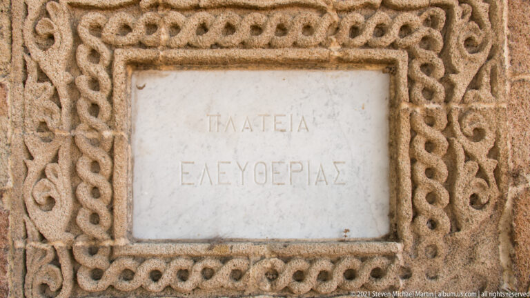 Greek stone plaque reading “ΠΛΑΤΕΙΑ ΕΛΕΥΘΕΡΙΑΣ” within an ornate looping carved border.