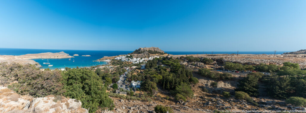 Wide coastal landscape near Lindos with a sheltered bay, distant white village, and fortified acropolis on a rocky hill.