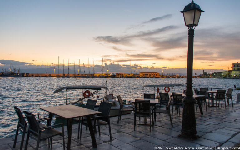 Outdoor café tables beside a harbor at sunset, with a tall streetlamp, rippled water, and boats beyond.