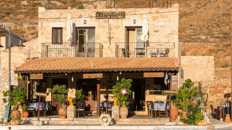 Two-story stone taverna with Greek signs, shaded outdoor tables, potted plants, balcony, and closed white umbrellas.