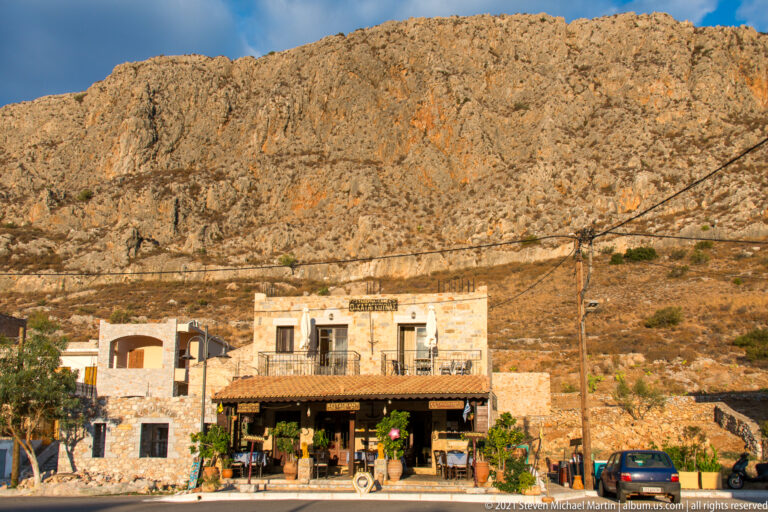 Small stone roadside taverna and parked car beneath a steep, barren rocky mountainside.