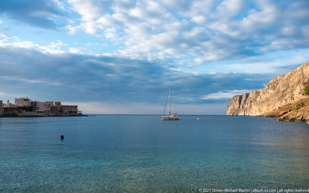 Sailboat anchored in a calm blue bay between a low waterfront village and a steep pale rocky headland.