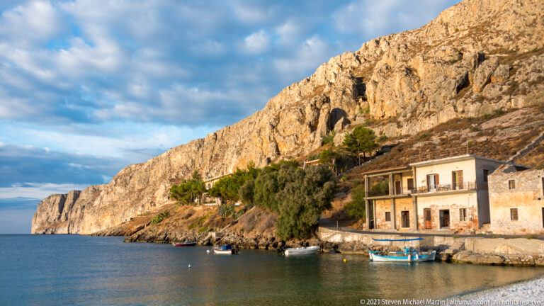 Stone waterfront houses and a small boat squeezed between calm sea and a towering rocky cliff.