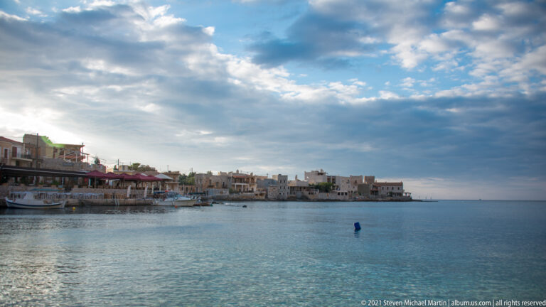 Low waterfront village across calm blue water beneath layered evening clouds, with terraces extending along the left shore.