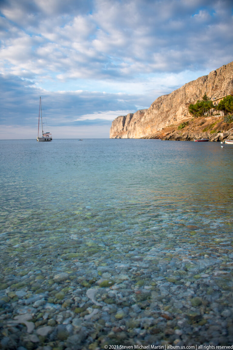Anchored sailboat beyond clear pebble-filled shallows, with a low rocky headland extending into the sea at right.