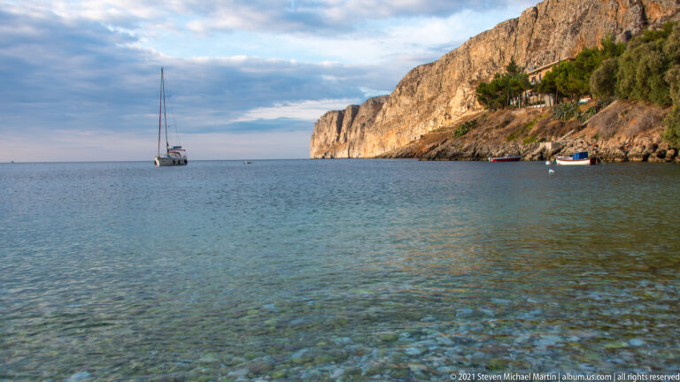 Anchored sailboat in clear water beside a steep pale cliff, with a smaller boat near the rocky shoreline.