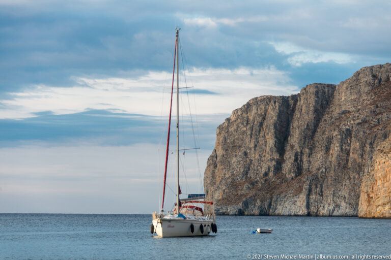 White cruising sailboat with tall mast anchored in front of a sheer gray-beige coastal cliff.