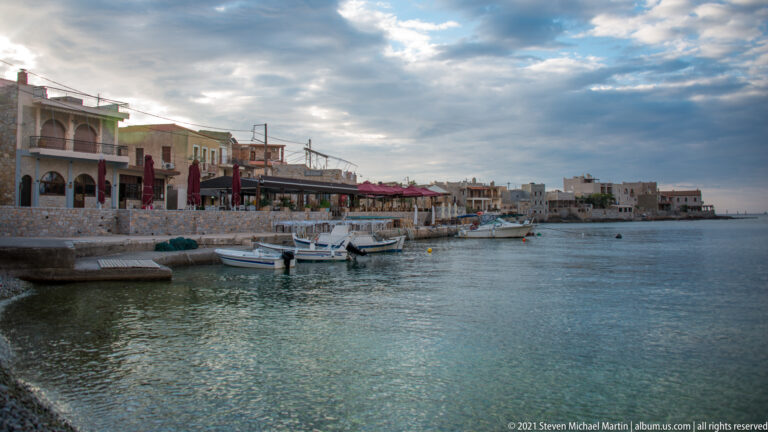 Small boats moored beside a stone quay lined with cafés and low waterfront buildings under a cloudy sky.