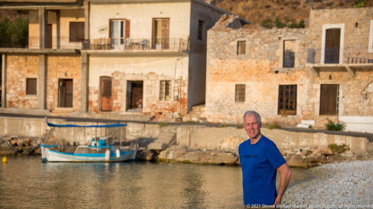 Man in a blue shirt standing by calm harbor water, with a small boat and old stone buildings behind.