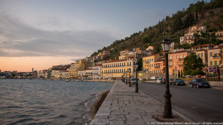 Stone waterfront promenade beside colorful buildings and a road, with houses rising on a steep hillside above.
