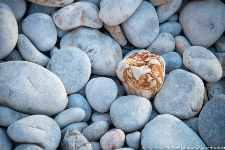 Close-up of smooth gray beach pebbles surrounding a white stone streaked with rusty orange veins.