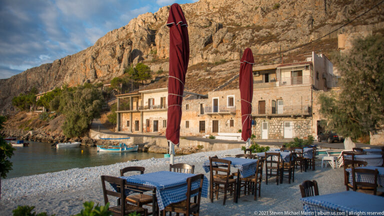 Seaside dining tables with blue-checked cloths and closed red umbrellas before stone houses and a steep cliff.