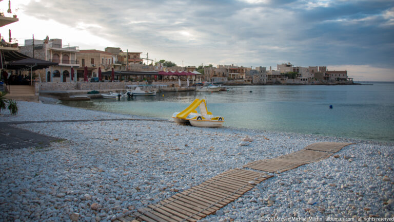 Wooden walkway across a white-pebble beach toward calm water, with a yellow pedal boat and village beyond.