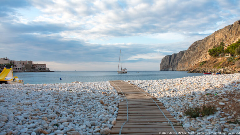 Wooden walkway crosses pale pebbles toward a sailboat in the bay, with cliff at right and village at left.