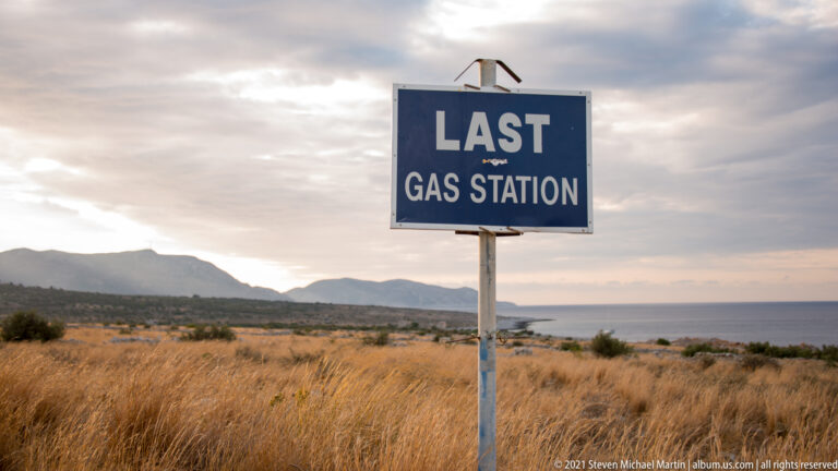 Blue roadside sign reading “LAST GAS STATION” above dry grasses, low mountains, and distant sea.
