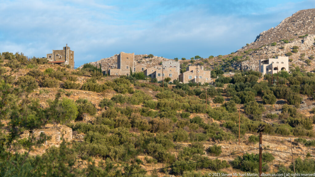 Scattered stone houses and tower-like buildings across a dry terraced hillside with shrubs and rocky slopes.
