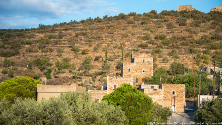 Tall stone tower house above lower buildings and trees, with additional towers scattered across the dry hillside behind.