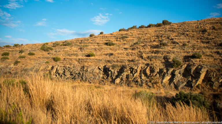 Dry hillside crossed by horizontal stone terraces, with golden grasses and exposed dark rock in the foreground.