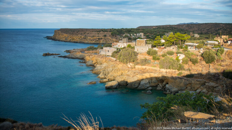 Stone houses clustered beside a clear blue rocky cove, surrounded by scrub and low coastal hills.
