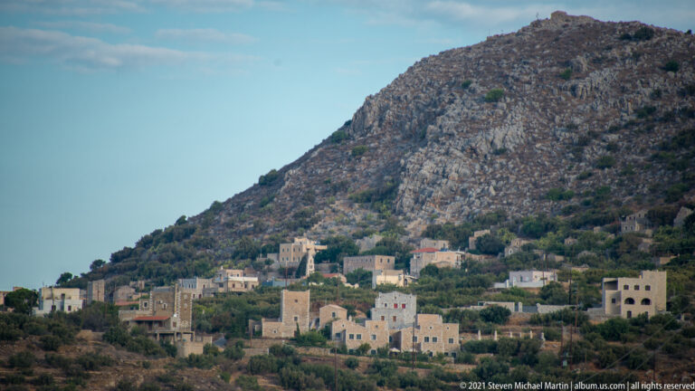 Pale stone village with tower-like buildings at the foot of a steep rocky hill topped by a small structure.