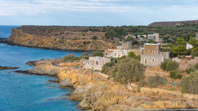 Stone houses and a tower-like residence beside a rugged ochre shoreline and deep blue coastal water.