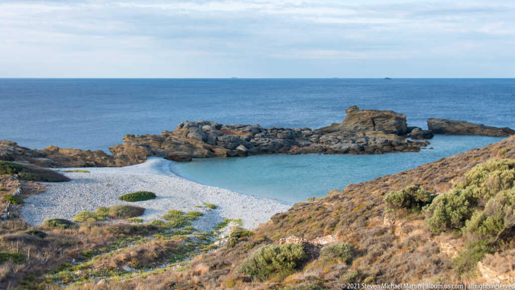 Rocky cove with clear blue water, pale shoreline, scrub-covered slopes, and open sea beyond.
