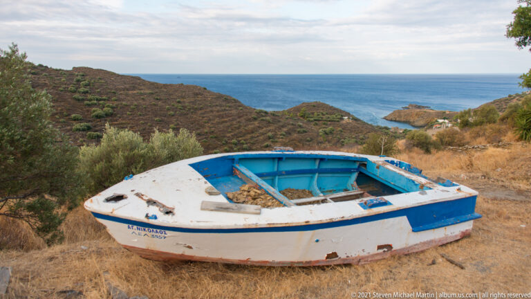 Blue-and-white wooden boat resting on dry grass above a distant rocky coast and sea.