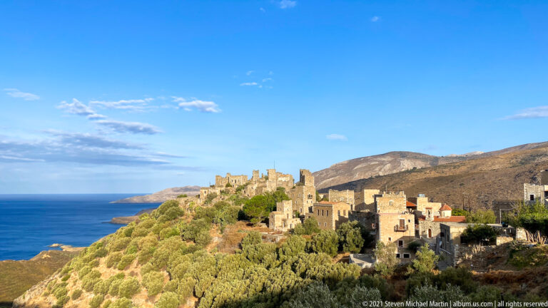 Stone tower-house village on a ridge above the sea, backed by dry mountains.