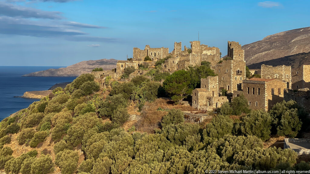 Sunlit stone tower houses clustered on a vegetated coastal slope with sea visible at left.