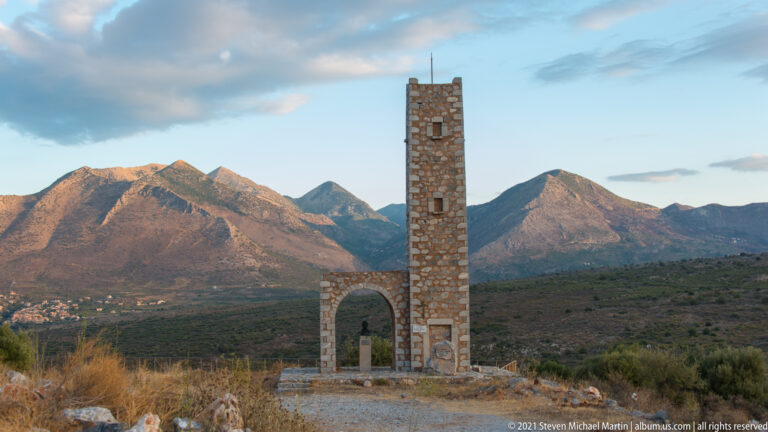 Tall narrow stone tower with an arched base and small openings standing before rugged sunlit mountains.