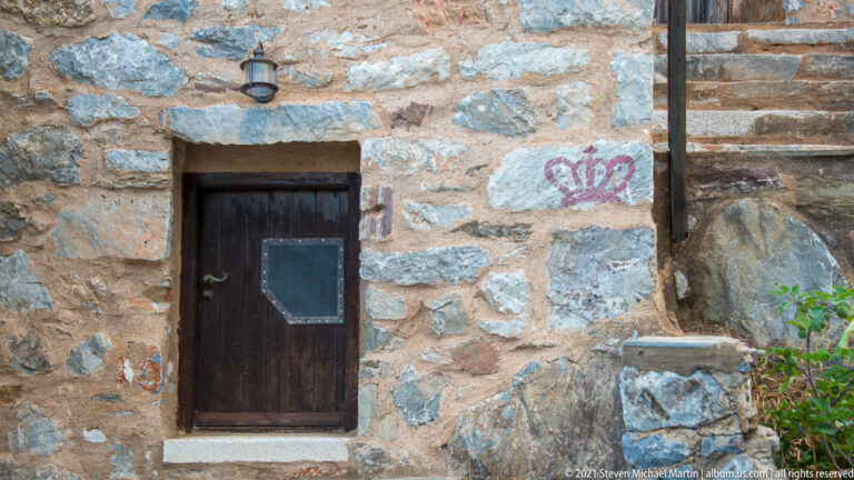 Dark wooden door in a rough stone wall beside a red painted crown-like symbol and exterior steps.