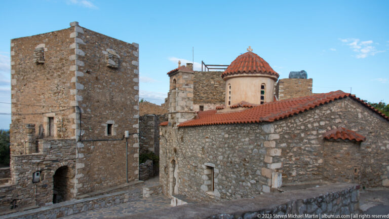 Small stone church with red-tiled dome beside tall rectangular tower houses in a Mani village.