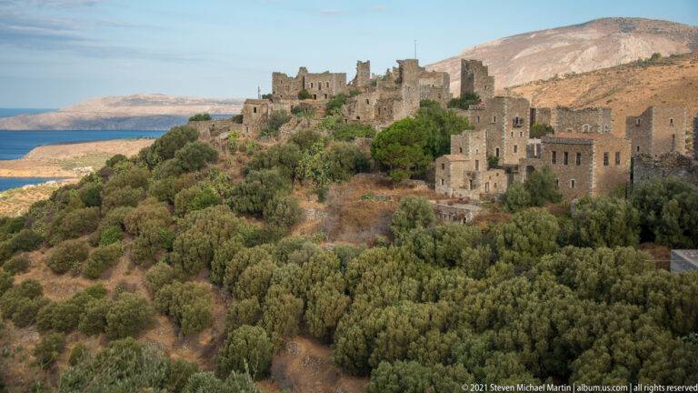 Stone tower-house village on a vegetated ridge above the sea with mountains behind.