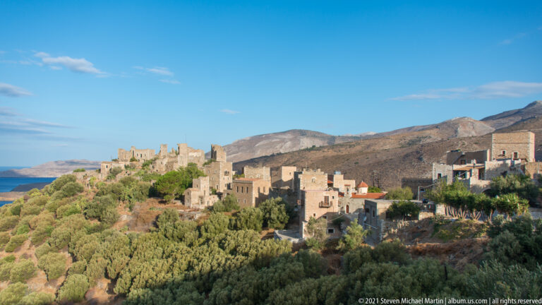 Coastal Mani settlement with stone towers, newer buildings, sea at left, and barren mountains behind.
