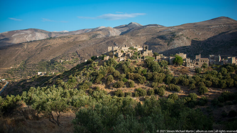 Small stone village on a green-brown hill beneath broad barren mountains and deep afternoon shadows.