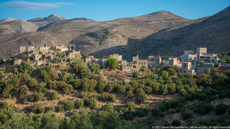 Stone village extending across a wooded ridge below high, sparsely vegetated mountains.