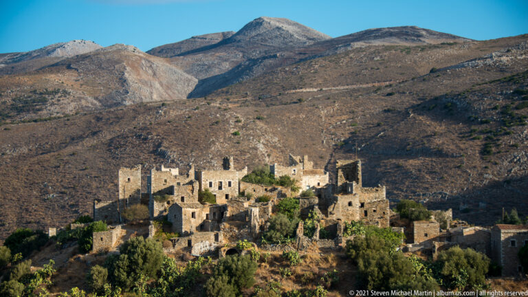 Sunlit stone tower-house village at the base of steep barren mountains, with deep foreground shadow.