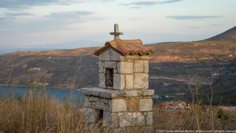 Small stone wayside shrine with red-tiled roof and cross, standing in dry grass above a coastal valley.