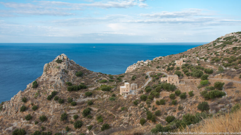 Dry rocky hillside with scattered stone buildings descending toward a pointed headland and blue sea.