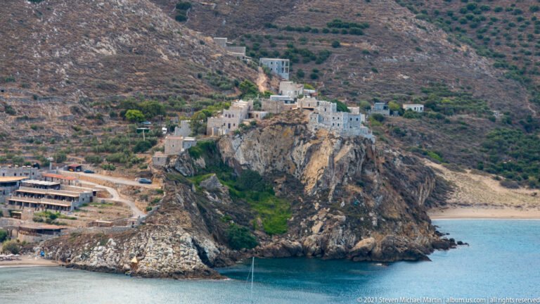 Pale buildings perched on a steep coastal rock above a small blue bay and shoreline road.
