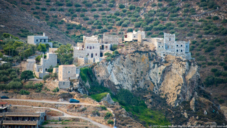 Pale buildings crowded atop a sheer gray coastal rock with road and vegetation below.