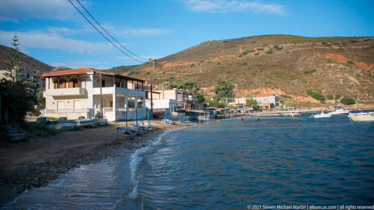 Waterfront houses lining a narrow pebbly shore beside calm blue water and dry hills.