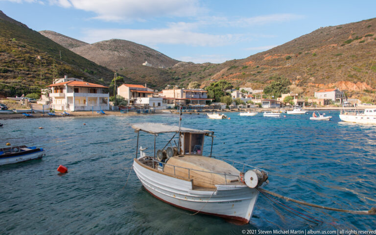Small open motorboat moored in calm harbor water with village houses and other boats behind.