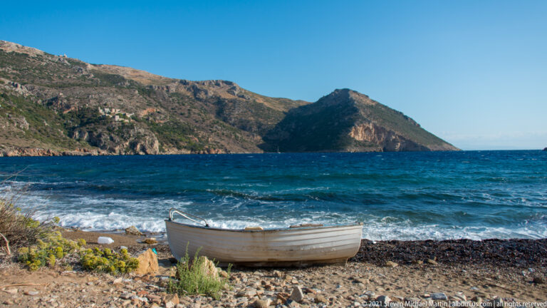 Small pale boat resting on a rocky beach with breaking waves and a rugged headland beyond.