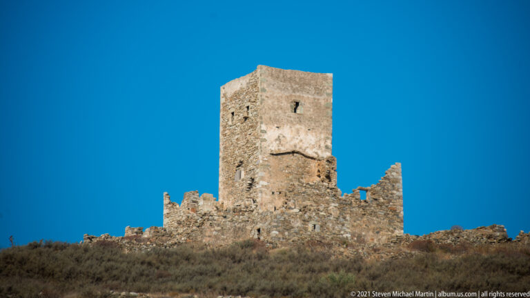 Tall rectangular stone tower rising above low ruined walls on a dry hillside under deep blue sky.