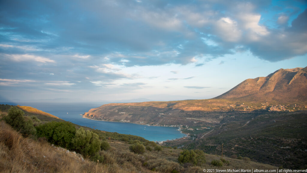 Blue bay curving below rugged mountains and a small hillside settlement, viewed from a dry scrub-covered slope.