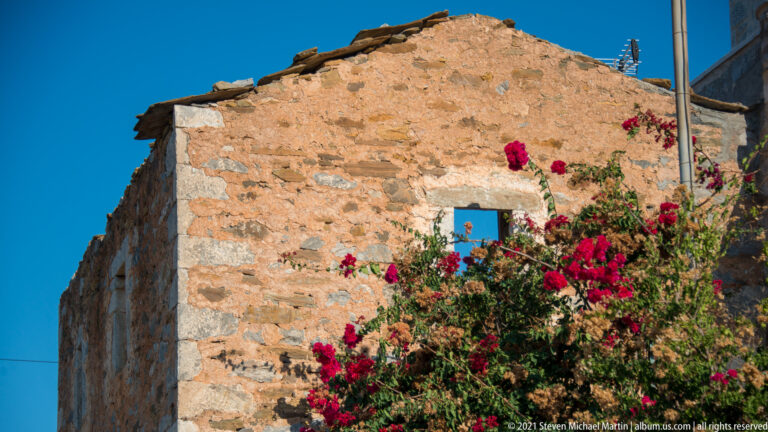 Weathered stone wall with a small square opening and bright bougainvillea against blue sky.