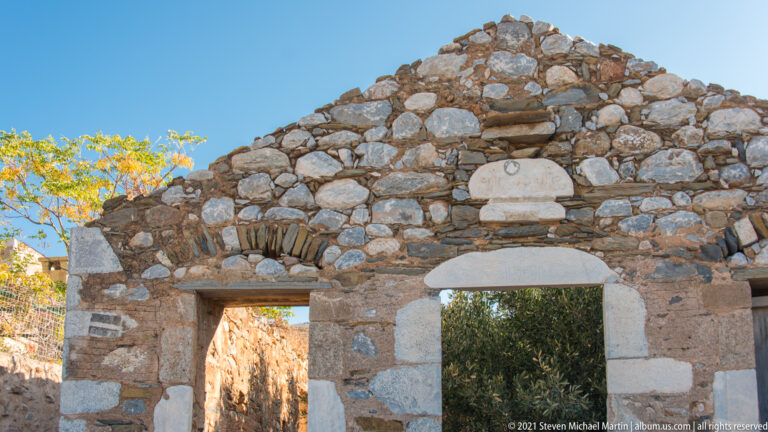 Rubble-stone chapel façade with arched openings, pale stone doorway, and marble plaque beneath the gable.