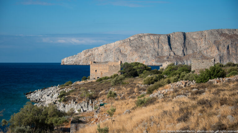 Stone fortified building on a rocky shoreline with a high pale limestone headland across the water.